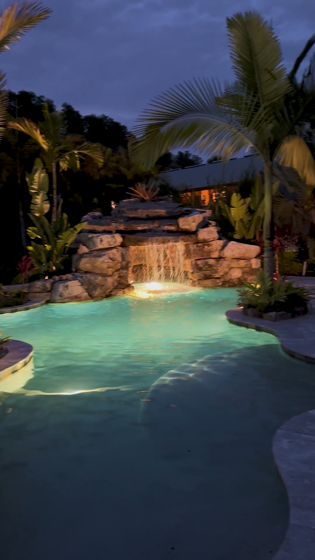 This natural rock grotto waterfall features a huge cave that can accommodate twelve people inside! We designed lush landscaping with lots of curing lines that blend the turf, tile, and beach pebbles throughout this tropical oasis located in Lutz, just outside Tampa, Florida. #insanepools #lucaslagoons #pool #poolbuilders #backyard #luxurylife 