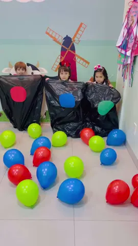 Essa dinâmica envolve crianças brincando em uma atividade divertida, que parece ser uma corrida de saco ou uma gincana com balões. As crianças estão dentro de grandes sacos pretos, provavelmente adaptados como fantasias ou obstáculos para a brincadeira. Elas seguram ou tentam pegar balões coloridos que estão espalhados no chão, criando um ambiente animado e alegre. O fundo da sala é decorado com imagens lúdicas, como uma vaquinha e um moinho de vento, o que reforça o ambiente infantil e descontraído. Essa atividade incentiva a coordenação motora, a interação social e proporciona uma experiência de aprendizado por meio de jogos.