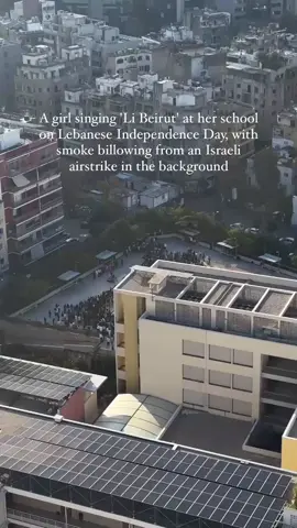 A girl singing ‘Li Beirut’ at her school on Lebanese Independence Day, with smoke billowing from an Israeli airstrike in the background.❤️‍🩹#lebanon #beirut #foryou 