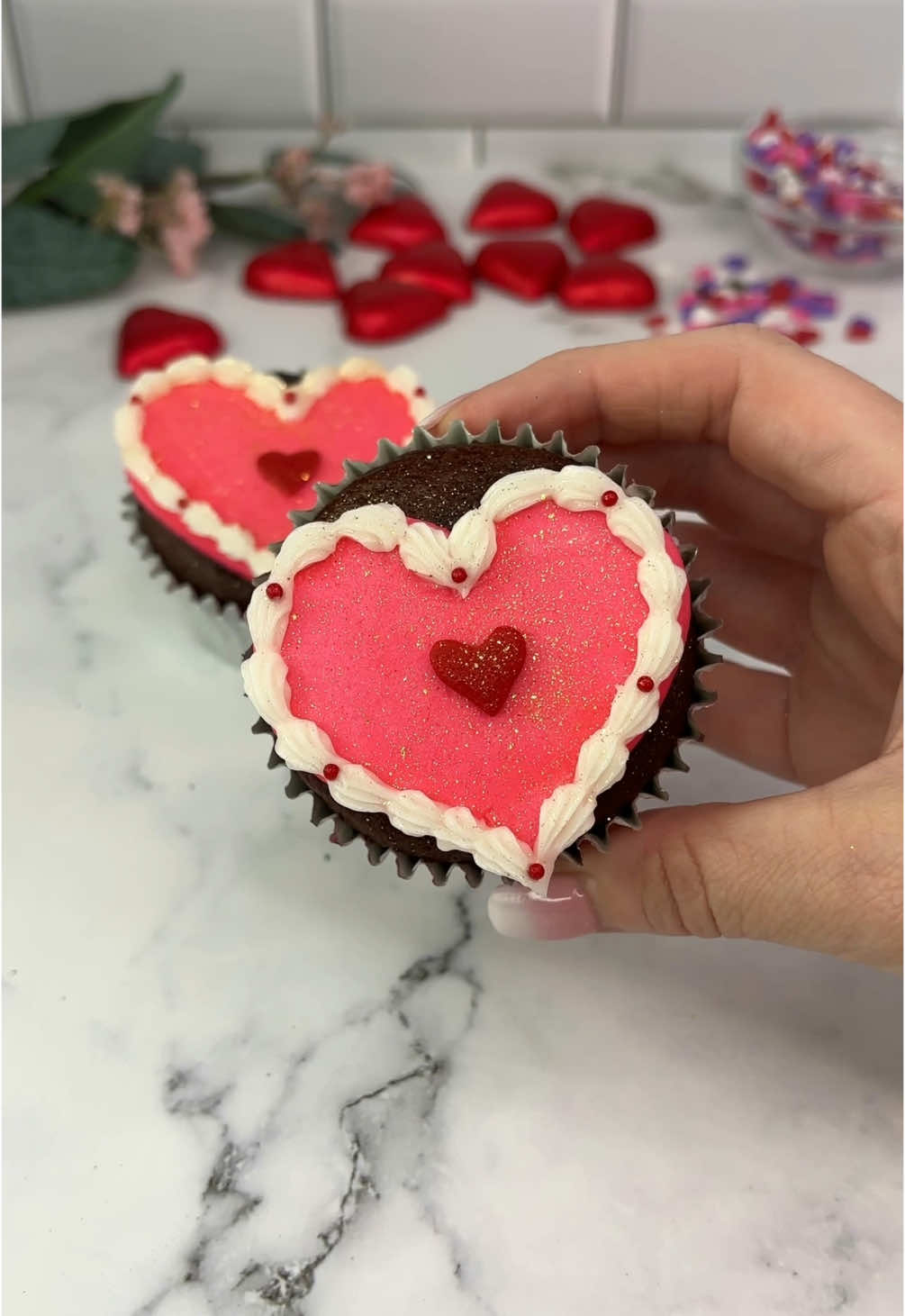 Buttercream heart cupcakes 💕🧁 💕 Use a large round piping tip to pipe a heart shape on the top of your cupcake.  💕 Smooth it out. 💕 Sit is gently onto parchment paper. You don’t want to loose the shape of the heart here, just to get a perfectly smooth finish.  💕 FREEZE FOR 15 MINUTES. 💕 Peel away from the parchment and decorate as you like.  💕 I added a border using tip 18 and then added a few sprinkles and a candy heart.  💕 I added some gold edible glitter ✨ I actually wish that I didn’t add this as I preferred them before. Once added it’s not coming off 🫣😝 #cupcakes #cakedbyrach #valentinesbaking #valentines #heartcake #heartcupcakes #cakeart #cakedecorating  