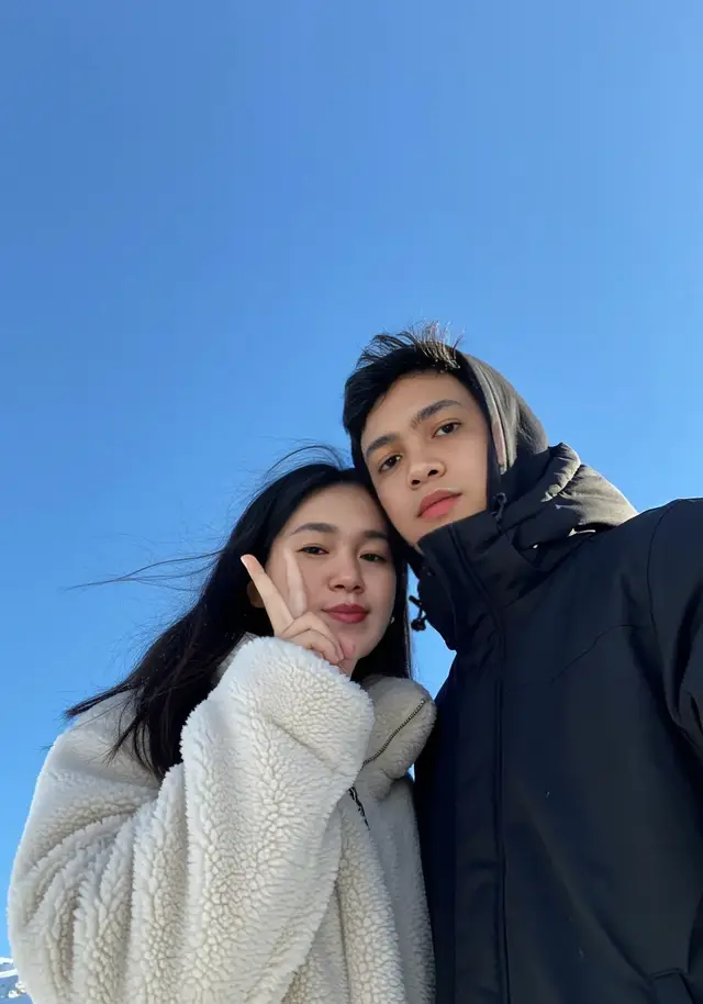 Prompt: Sebuah selfie pasangan yang diambil dari sudut bawah (low angle) dengan latar langit biru cerah tanpa awan. Kedua orang dalam foto — pria dan wanita — tidak memakai helm, hanya mengenakan kacamata ski besar berwarna pelangi (rainbow reflective lens) yang memantulkan cahaya matahari dengan warna-warni yang indah. Pria berada di sisi kanan frame, mengenakan jaket musim dingin warna hitam doff dengan hoodie turun. Wajahnya tertutup sebagian oleh kacamata ski pelangi, namun senyumnya terlihat jelas. Ia memegang kamera dengan tangan kiri, menciptakan komposisi selfie dari bawah. Pacarnya berdiri di sisi kiri frame, lebih condong ke arah kamera. Ia terlihat sangat cantik dengan jaket puffer warna putih tulang (off-white) yang fluffy, dan kacamata ski pelangi besar yang menutupi separuh wajahnya. Rambut panjangnya terurai bebas, tidak ditutup helm, dan beberapa helai tertiup angin musim dingin. Dia membuat pose lucu: duck face atau bibir sedikit maju, dan tangan kirinya membentuk tanda peace (✌️) di dekat wajah. Pipinya tampak merah muda alami karena cuaca dingin. Kedua orang tampak sangat akrab dan bahagia, berdiri sangat dekat satu sama lain. Bayangan ringan dari cahaya matahari sore memberikan efek lembut di wajah mereka. Latar belakang didominasi oleh langit biru jernih, memberikan kontras sempurna dengan warna kacamata pelangi mereka dan jaket-jaket tebal musim dingin. Salju putih tampak di bagian bawah frame sebagai aksen alami. Suasana keseluruhan sangat ceria, hangat, romantis, dan penuh cinta, khas liburan musim dingin berdua. #couplegoals #AI #chatgpt #outfitideas #jakarta #indonesia🇮🇩 #trending #tiktok #foryoupage 