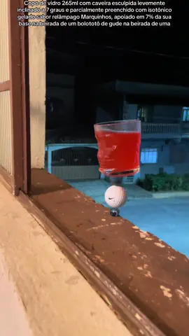 Copo de vidro 265ml com caveira esculpida levemente inclinado a 7 graus e parcialmente preenchido com isotônico gelado sabor relâmpago Marquinhos, apoiado em 7% da sua base na beirada de um bolototô de gude na beirada #coposnabeirada #coponabeirada #copo #beirada 