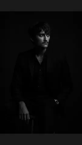 A dramatic black and white portrait of a confident man sitting on a wooden stool against a dark studio background. He is wearing a well-fitted, all-black suit with a black shirt, and black pant small silver chain at naki and small silver watch slightly unbuttoned at the top, exuding a moody and powerful aura. His posture is relaxed yet dominant, with one arm resting on his leg and the other one in his pant pocket. The lighting is soft but directional creating.#trending #fyp #ai #img #chatgptimage #chatgpt 