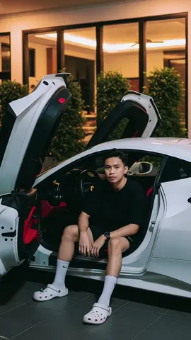 Seorang remaja pria duduk santai di dalam mobil buatkan saya foto sport mewah Ferrari berwarna putih dengan pintu terbuka model gullwing. Ia mengenakan kaos hitam, celana pendek hitam, kaus kaki putih tinggi, dan sandal Crocs putih. Ia berpose dengan gaya tangan damai (peace sign) sambil menatap kamera. Interior mobil bernuansa hitam dan merah. Lokasi berada di garasi rumah modern di malam hari, dengan pencahayaan hangat dan latar belakang berupa jendela besar serta tanaman hijau terpangkas rapi. Suasana foto bergaya candid dan mewah. #editfoto #chatgpt #geminiai #fyppppppppppppppppppppppp 