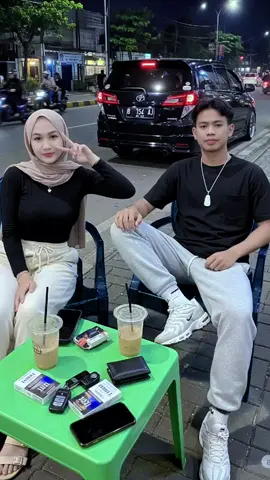 lagi mager tapi pengen punya pasangan ketik aja:A young couple sitting casually on a city sidewalk at night, at a roadside coffee stall. The man wears a black t-shirt, light gray jogger pants, and white Nike sneakers, with a white necklace around his neck. The woman wears a light brown hijab, black long-sleeve top, cream-colored pants, and casual sandals. In front of them is a green plastic table with two iced coffee cups, cigarettes, wallets, car keys, and phones. The background shows a lively urban street with passing cars and motorcycles, glowing street lights, and a relaxed evening vibe. Realistic photo style, candid street atmosphere, high quality, 4K. #fypppppppppppppp #editfoto #googelveo3 #geminiai #chatgpt 