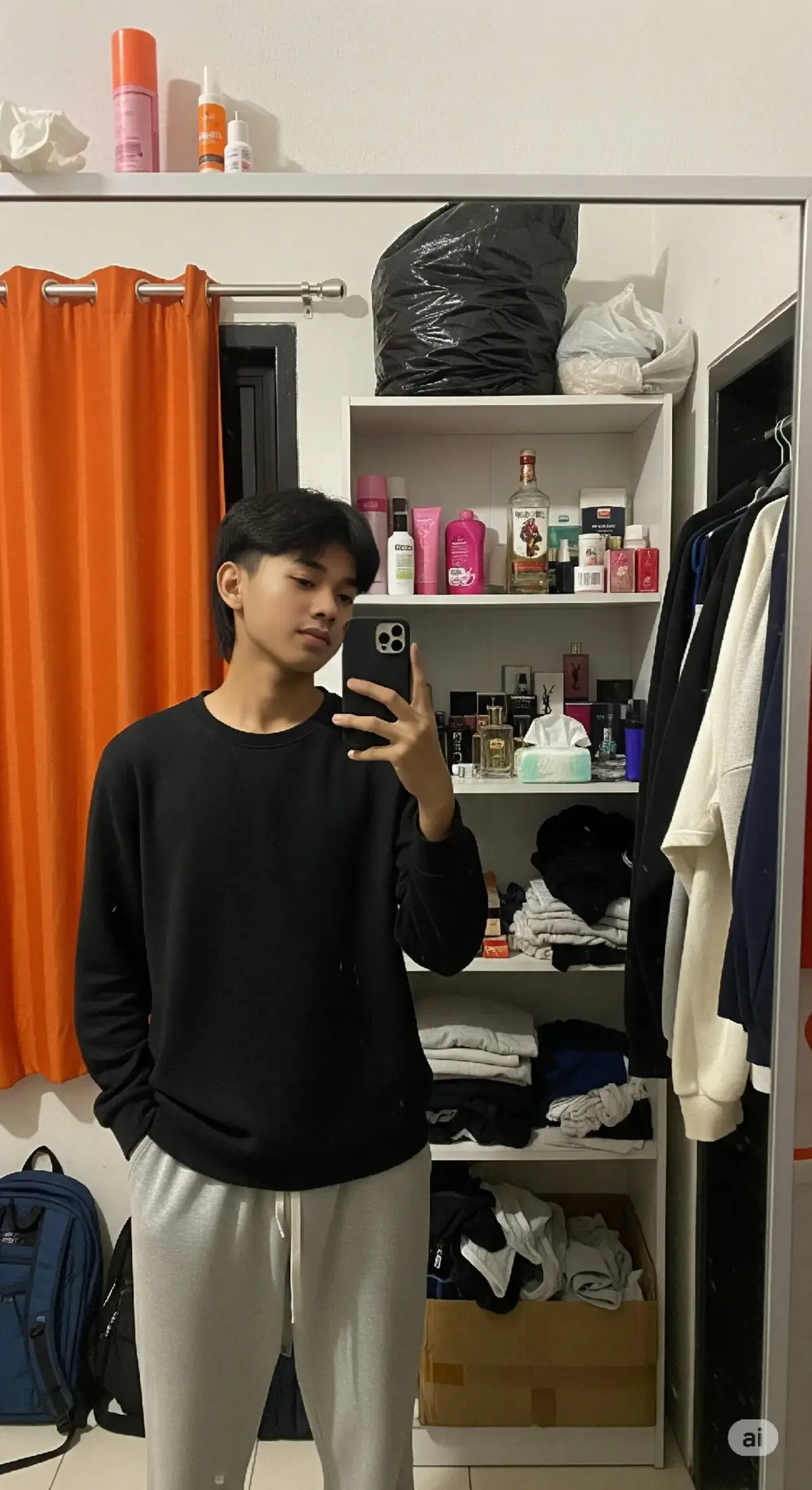 Seorang remaja laki-laki berwajah indonesia berumur 17 tahun, tinggi badan 167 cm berat badan 47 berdiri di depan cermin besar untuk mengambil foto selfie menggunakan ponsel. Ia berdiri sedikit menyamping dengan tangan kanan memegang iPhone 13 yang diarahkan ke cermin. Wajahnya berkulit cerah dengan ekspresi datar dan percaya diri, rambutnya hitam lurus ke belakang dengan poni tepat di atas mata. Ia mengenakan sweater lengan panjang warna hitam polos yang pas di tubuh bagian atas dan celana training abu-abu muda yang longgar, menjuntai hingga ke bawah. Cermin yang digunakan memiliki bercak debu dan noda air, menunjukkan bahwa cermin jarang dibersihkan. Cermin bersandar di dinding kanan ruangan, bersebelahan dengan jendela berbingkai hitam yang tertutup sebagian oleh tirai berwarna oranye terang. Di depan jendela, di atas permukaan dinding putih, terlihat sebuah lip balm atau salep bibir warna oranye dan lip balm tube warna pink yang tergeletak. dan tisu bekas Di belakang remaja tersebut terdapat sebuah lemari rak terbuka berwarna putih dengan beberapa barang pribadi di dalamnya. Di rak kiri atas, ada plastik besar berwarna hitam berisi barang yang tidak terlihat jelas. Di rak kiri bawah, terlihat dua ransel: satu warna biru tua dan satu lagi hitam, tergantung ke bawah dengan tali menjuntai. Rak kanan atas diisi oleh berbagai produk perawatan tubuh dan rambut: terlihat botol pink yang mungkin body lotion, spray botol pink transparan, botol-botol kecil warna-warni, juga botol captain morgan dan koleksi parfum YSL dan beberapa kotak kecil. Ada juga tisu atau lap di dekatnya. Rak kanan bawah terlihat agak berantakan dengan pakaian yang dilipat asal-asalan, serta barang-barang acak yang sulit dikenali khas kamar indonesia. Bagian bawah lemari (lantai) terlihat terdapat beberapa kotak kardus dan barang-barang lain yang menumpuk, seperti buku atau map warna gelap, memperkuat kesan bahwa ruang ini digunakan sehari-hari dan tidak terlalu rapi. dan celana dalam acak acakan Di sisi kanan luar cermin tergantung beberapa potong baju berwarna hitam, putih, krem, dan navy. Satu di antaranya tampaknya adalah jaket atau hoodie. Pencahayaan ruangan berasal dari lampu indoor dengan cahaya putih kekuningan yang membuat suasana terlihat hangat dan nyaman, meskipun ruangannya cukup padat dan penuh barang. rasio 9.16 #prompt #ai #aigemini #trend #fyp 
