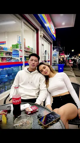 PROMPT? ORDER? DM A night scene outside the typical red-blue-yellow Indomaret. A man and a woman sit in casual poses, only visible from the waist up (close-up). The man is seated casually in a black metal chair with a black round table partially visible in the frame. On the table are several items: a white iPhone, a lit cigarette in the ashtray, a pack of cigarettes, a Coca-Cola drink, and snacks. The background shows the Indomaret's large glass windows reflecting light from the lights, shelves visible from inside, neatly arranged gallon jugs of mineral water on the left side, a round aluminum trash can, and silhouettes of activity such as shoppers, motorcycle taxi drivers driving through the drive-through, parked vehicles, young people hanging out, and other outlets in the distance. Asian woman with oval face, long wavy hair, wearing a black crop top and shorts. She is seen leaning toward the male model with a happy expression. Asian man with white face, oval face, black quiff hair, wearing a ripped denim jacket and long black cargo pants. He looks at the camera with a happy expression. (No bokeh), no AI artifacts, detailed and realistic skin textures, like those taken with a phone camera, natural lighting and shadows, accurate human body proportions, realistic fabric folds, and depth of field. A vibrant and natural atmosphere, like a real photo. Aspect ratio: 3:4 #ai #gemini #geminiairsign #chatgpt #aifaceswap #MLBBSkinGifts #freefire #devinakaramoy #faceawapapp 