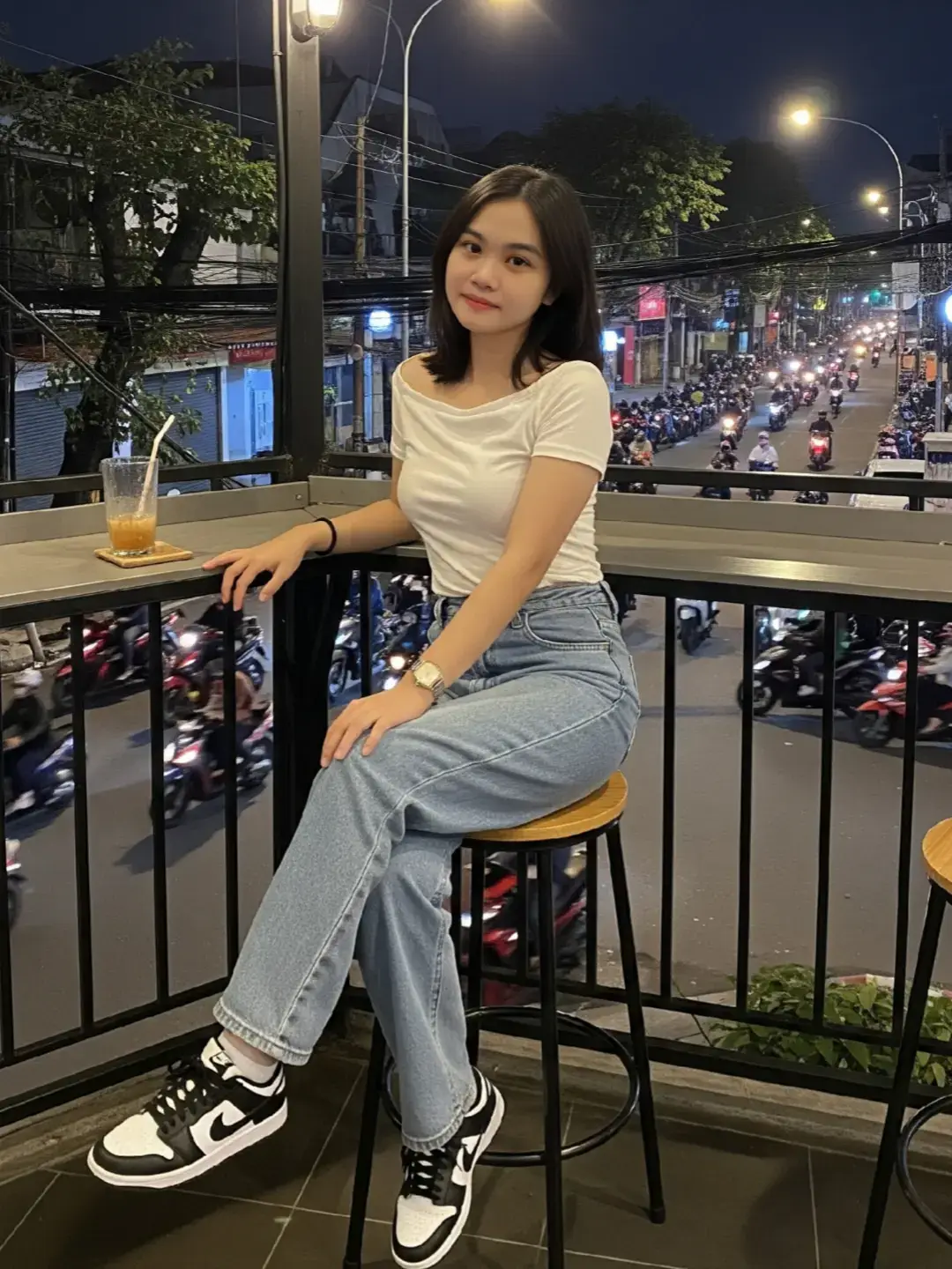 A young indonesian woman sitting casually on a high stool at an outdoor balcony cafe at night, with a busy street full of motorbikes in the background. She is 21 years old, with fair skin bright, her blackish brown hair, arm length, height 156cm, weight 50cm, oval face shape with thin cheeks and sharp chin. She has black hair along the arm, wears a stylish black crop top strict (with the chest slightly open), loose high-waisted blue jeans, and black-and-white Nike sneakers. She is holding a drink with one hand resting on the counter the other hand holds the thigh while crossing the legs gracefully, the body is straight facing the camera, looking at the camera with a soft expression. The setting is urban, with warm cafe lighting and nighttime street lights. (whole body straight facing the camera). #prompt #AI #4upage #gemini #chatgpt