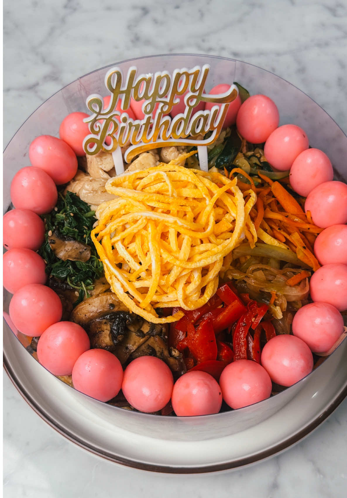 healthy japcake 😆🎂 #alexcooks  merangkap 3 in 1: mie ultah, kue dan sehat HEHEHE jujur seru banget bikin iniii merasa tertantang dan hasilnya memuaskan bgt! total untuk porsi 6-8 orang minyak yang kepake ga nyampe 3 sdm 🥹 dan manis juga pake stevia jadi lebih aman. yang terpenting: rasa tetep enak pol walaupun sehat~ Bahan: 300 gr mie shirataki kering 1 bombay 1 paprika merah 1 wortel ukuran besar 1 zukini 7 jamur champignon 2 telur 30 telur puyuh (opsional) pewarna pink biji wijen secukupnya garam secukupnya lada hitam secukupnya Minyak olesan: 1/2 sdt minyak goreng 1 sdt minyak wijen Bayam: 1 ikat bayam, cuci bersih 1/3 sdt garam 1/4 sdt lada hitam 1/2 sdt minyak wijen 1/2 sdt biji wijen Ayam: 2 dada ayam (250 gr) 1 1/2 sdt garam 1/2 sdt gula (opsional) Total 400 ml air  2 bawang putih, cincang 1 sdm kecap asin 1 tetes stevia 1/2 sdt minyak wijen 1/3 sdt lada hitam Bumbu: 10 sdm kecap asin 9 tetes stevia 1/3 sdt micin 2/3 sdt kaldu bubuk Cara membuat: 1. Ayam: Larutkan garam dan gula dengan sedikit air panas. Tuang sisa air dingin lalu masukkan dada ayam. Pastikan ayam terendam sepenuhnya lalu diamkan di kulkas selama 1 jam. Keluarkan dan keringkan. Iris tipis, masukkan bumbu dan bawang putih, aduk rata dan marinasi di kulkas minimal 30 menit. 2. Oles sedikit minyak di pan antilengket lalu masukkan bombay dengan sedikit garam dan lada. Masak di api sedang hingga matang. Ulangi proses yang sama ke zukini, wortel dan paprika. Boleh tambah sedikit air sedikit ke pan bila terlalu kering atau pan terlalu panas. 3. Masukkan jamur bersama sedikit air ke pan lalu masak hingga jamur menciut dan air menguap. Masukkan garam, lada dan sedikit minyak wijen lalu aduk rata. 4. Kocok telur, dadar, tunggu dingin lalu iris tipis. 5. Rebus telur puyuh selama 7 menit lalu rendam di air es. Kupas, tetes pewarna pink secukupnya dan aduk rata. 6. Rebus bayam selama semenit atau cukup hingga layu lalu angkat dan rendam di air es. Peras lalu potong sedikit. Masukkan sisa bumbu dan aduk rata. 7. Rebus mie shirataki kering sesuai petunjuk hingga matang lalu tiriskan. Saat masuh panas, tuang 1/3 bumbu dan biji wijen lalu aduk rata. 8. Masukkan bombay, paprika, wortel, zukini, bayam, jamur, dan ayam ke mie (sisakan sedikit untuk topping). Tuang sisa bumbu dan biji wijen lagi bila perlu, aduk rata. 9. Tuang ke cetakan. Tata telur dadar iris, sisa topping dan telur puyuh. Sajikan.