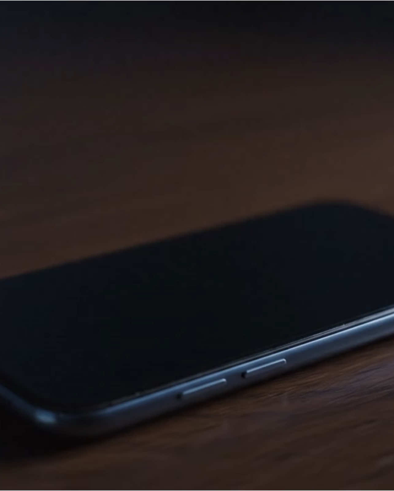 A cinematic 8-second video begins with a close-up of a smartphone resting on a wooden desk. The phone suddenly vibrates and receives a rapid flood of notifications. As the pressure builds, the screen visibly bulges outward. Suddenly, it shatters violently, releasing glowing notification icons and black glass shards that burst into the air. The phone lies broken as hundreds of physical notification symbols swirl above it in slow motion, casting shadows and sparks in a darkened room. video prompt details: 16:9, photorealistic, high-speed camera, moody lighting.
