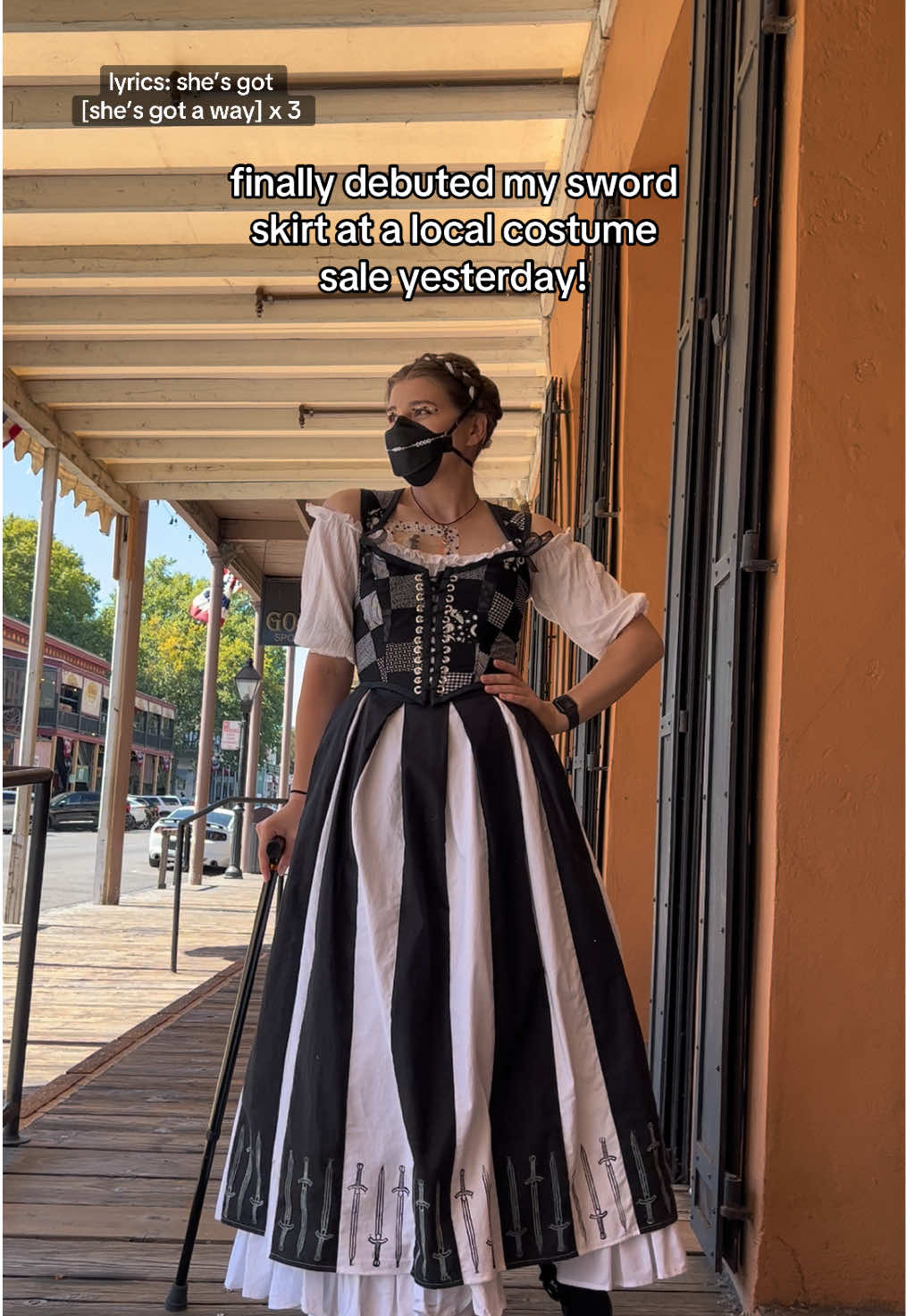 This outfit might be my final form (we love an entirely home made outfit) VD: Emma, a white thin young person with brown hair in a white ribbon braid crown, stands on a wood side walk with a porch overhang. They are wearing black and white patchwork stays, a black and white paneled skirt with swords at the hem, a white ruffled petticoat, a black N95 mask with a white mask chain, black heeled shoes, and are holding a black cane. Text reads “finally debuted my sword skirt at a local costume sale yesterday”  #CovidConscious #HighRiskCovid19 #Mask4Mask #ChronicIllness #Disability #SickTok #BabeWithAMobilityAid #SewistsOfTikTok #WhimsicalAesthetic #MeMadeWardrobe #HistoricalCostuming 