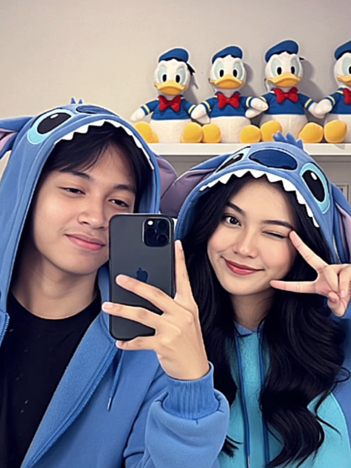 prompt Gemini 👇 Buatkan foto pantulan cermin besar realistis pasangan muda Indonesia dengan suasana ceria di dalam ruangan. Mereka mengenakan hoodie karakter Stitch berwarna biru lengkap dengan telinga dan wajah karakter di bagian kepala. Latar belakang terdapat boneka Donald Duck berjajar di rak. Wanita berumur 18 tahun di sebelah kanan berambut panjang hitam, wajah cantik, sedang tersenyum lembut sambil mengedipkan satu mata. Pria berumur 18 tahun di sebelah kiri berambut hitam lurus, mengenakan kemeja hitam muda, tersenyum tipis sambil memandang kamera. Mereka memegang ponsel hitam untuk selfie. Gaya fotonya menggunakan kamera iPhone 14 dengan kualitas resolusi agak rendah tidak terlalu HD, kontras rendah, bayangan lembut, tidak ada efek estetika tambahan, foto terlihat original tidak seperti di edit. Pencahayaan hangat, fokus tajam pada wajah, dan nuansa imut dan romantis. Rasio gambar 9:16.#prompt #jasaprompt #gemini #ldrstory #realistic 