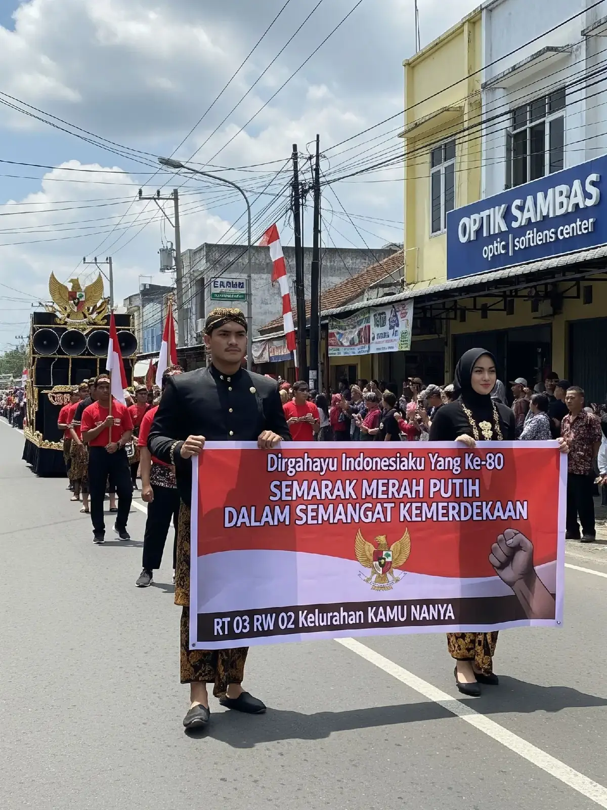 Promt Gemini: A photo of a sunny afternoon parade celebrating Indonesian Independence Day. The setting is a main road with a row of shophouses on the right side, one of which reads 'OPTIK SAMBAS optic | softlens center'. Power lines and poles are visible along the road. Spectators stand and sit on the roadside to watch the parade. The weather is sunny with a few white clouds, and clear shadows are visible on the asphalt. In the foreground, a man walks with a woman wearing a black hijab wrapped around her head. The man wears a plain black long shirt similar to a beskap, batik trousers with a parang motif, dark closed-toe shoes, and a gold/brown blangkon. The woman wears a simple but formal long-sleeved black shirt, and a batik sarong with a parang or kawung motif. They both hold a large red and white banner that says 'Happy 80th Indonesia' at the top, 'Red and White Vibration in the Spirit of Independence' as the main slogan, and 'RT 03 RW 02 Kelurahan KAMU NANYA' with an illustration of a clenched fist at the bottom. Behind them, other parade participants wore similar black costumes with traditional Javanese accents and carried the Indonesian flag, below which was the One Piece Jolly Roger flag and the Garuda emblem. A sparkling horeg sound system was also present, along with dancers wearing red shirts, black trousers, and shoes. 3:4 ratio #gemini #kemerdekaanindonesia #karnaval #onepiece #17agustus 
