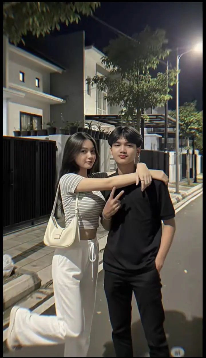 PROMPT : Sebuah foto yang sangat realistis diambil dari sudut kamera samping tentang pasangan muda yang berpose bersama di jalan pada malam hari. Gambar ini juga menampilkan latar belakang kota modern dengan bangunan putih dan sebuah pohon. Di sisi kiri gambar, seorang wanita muda dengan rambut hitam panjang dan tubuh ramping, mengenakan crop top bergaris dan celana putih, berdiri di trotoar dengan tas tangan putih di bahu. Ia menghadap ke arah pemirsa dan memandang mereka dengan senyum. Rambutnya diatur dengan gaya rambut pendek dan ia mengenakan kaus hitam dan celana hitam. Tubuhnya ramping dan ia memiliki senyum di wajahnya. Di sisi kanannya, seorang pria Asia berusia 20 tahun dengan rambut hitam pendek berdiri dengan lengan mengelilingi leher wanita. Ia tersenyum dan membuat tanda perdamaian dengan tangannya. Ia mengenakan kaus hitam dan celananya juga hitam. Latar belakang menampilkan jalan dengan tiang lampu dan beberapa pohon. Pencahayaan yang redup menciptakan suasana yang murung. #gemini #aiphotography #photolab #trending #editphoto 