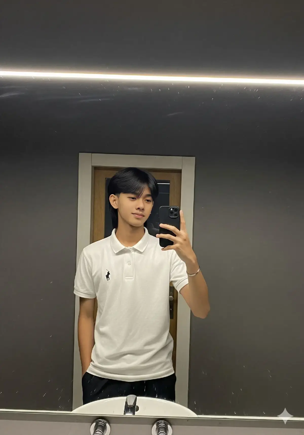 Buatkan foto seorang pria remaja asal Indonesia yang menarik sedang berfoto selfie di depan cermin sangat besar di dalam ruangan dengan dinding berwarna abu-abu gelap, kemungkinan sebuah kamar mandi mall. Pencahayaan terang memanjang horizontal di atas cermin, memantulkan cahaya ke bawah.  Pria tersebut menghadap ke cermin dengan sedikit memiringkan tubuhnya ke arah cermin. Ia memiliki tubuh kurus, bentuk wajah oval dan tatapan mata yang tajam.  Rambut hitamnya lurus agak panjang bergaya mullet dengan bagian poni belah tengah pas di atas mata. Ia juga mengenakan kaus polo putih polos dengan logo hitam kecil di bagian dada. Celana pendek hitam juga terlihat samar di bawah kausnya. Ia mengenakan gelang titanium di pergelangan tangan kirinya. Ekspresi santai dan sedikit tersenyum. Di bagian bawah cermin, tampak sedikit wastafel berwarna putih dan perlengkapan kamar mandi. Fokus utama gambar adalah seorang remaja dengan pakaian kasual.  Foto di ambil menggunakan kamera iphone 13 dengan kesan estetika khas kamera iphone,cahay lampu flash atau senter bagian belakang kamera menyala,resolusi tidak terlalu tinggi, kontras rendah,tanpa efek bokeh atau blur, belum di edit.  Rasio aspek 3:4 #prompt #googlegemini #chatgpt #Ai #fyp 