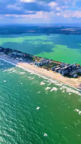 Do you ever crave a place that shouldn’t exist, yet somehow does? From above, Mamaia isn’t just a resort—it’s a fever dream stitched onto a blade of sand, a defiant sliver wedged between the restless Black Sea and the languid, mirror-eyed Lake Siutghiol. It’s the kind of geography you’d expect in a half-forgotten myth: too narrow to belong to the real world, too audacious to be accidental, as if the land itself decided it wanted to flirt with both water and sky at the same time. Here, the strip is a tightrope of pleasure, a ribbon of gold stretched between two liquid worlds with no room for hesitation. To the east, the sea crashes like an uninvited lover—loud, salty, insistent. To the west, the lake reclines in decadent stillness, holding the sun’s last bruised colors long after the day has given up. Above it all, the summer haze turns every rooftop, every beach umbrella, every human silhouette into part of a single living organism—restless, glowing, insatiable. Maybe that’s Mamaia’s real seduction: the feeling that you’re suspended between opposites, belonging wholly to neither. Sea or lake? Motion or stillness? Ferocity or surrender? The strip doesn’t care which you choose—it only dares you to walk it without looking down. So, which side would call you first? And would you ever cross to the other? Video by @pilot.mihai  [Black Sea Beaches, Mamaia Strip, Lake Siutghiol, Romanian Seaside, Coastal Aerial View, Narrow Landform, Romanian Riviera, Summer Destination, Beach Resorts, Coastal Geography, Seaside Escape, Dual Water Views, Coastal Paradise, Romanian Tourism, Water and Sand, Seaside Strip, Summer Travel, Beach Holidays, Eastern Europe Coast, Romanian Coastline] #romania #travel #beach #Summer 
