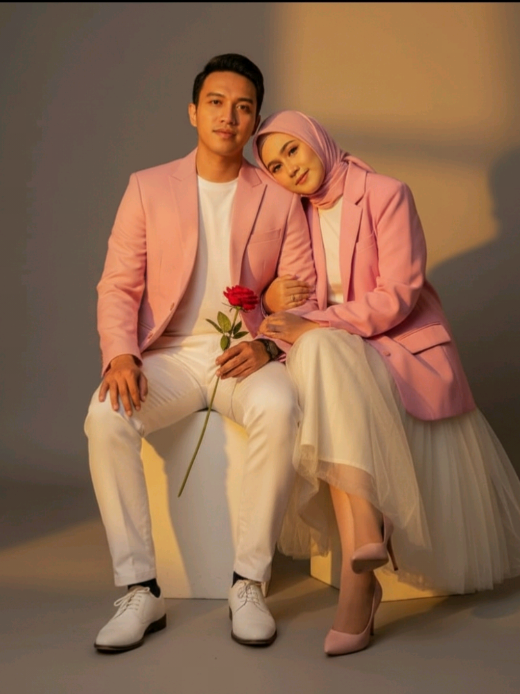 Edit foto ini menjadi pasangan duduk santai di studio foto dengan pantulan cahaya senja. Si cewek mengenakan outfit serba pink blazer oversize, dan rok tutu putih tulang high heels pink mewah, jilbab pink dan si cowok mengenakan outfit serba pink blazer dan celana putih, sepatu fantofel putih. Angel dari depan dan si cewek bersandar di bahu si cowok. Gaya elegan dengan dan edgy dengan nuansa misterius. memegang setangkai bunga mawar tanpa merubah wajah #fyp #gemini #geminiai #xyzbca #CapCut 