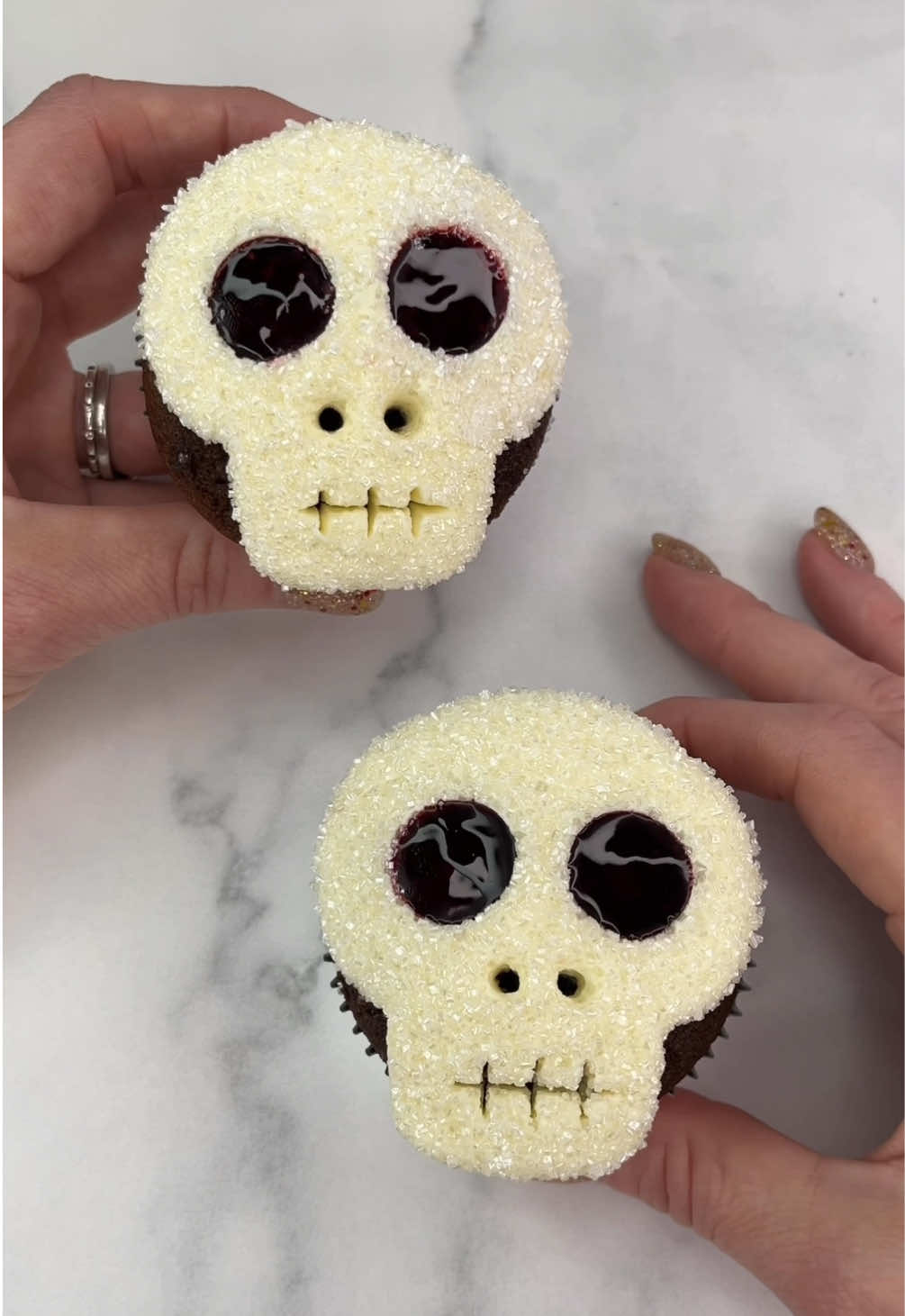 Sparkly Skulls 💀💀 Spooky, sparkly and so simple to make 💀🦴🧁 💀 Use white buttercream and piping tip 2A to pipe a skull shape onto your cupcakes.  💀 Smooth out any lines in the buttercream with a small spatula. 💀 Gently press them into white sanding sugar. 💀 Pop them in the fridge to firm up the buttercream.  💀 Use a piping tip to make holes for the eyes and nostrils.  💀 Use a toothpick to carve a mouth.  🩸Fill the eyes 👀 with strawberry sauce. #cupcake #cakedecorating #cutebaking #cakedbyrach #halloweenbaking   
