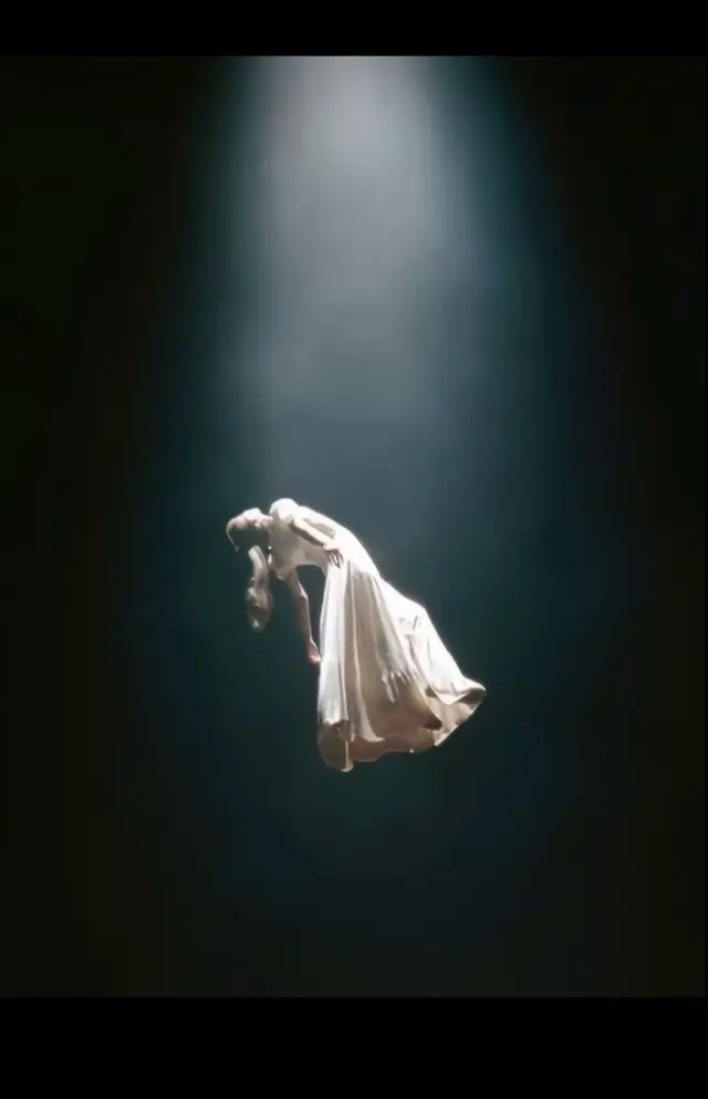 A surreal cinematic photo of a person floating weightlessly in mid-air under a single dramatic spotlight, surrounded by vast darkness. The figure appears to be gently suspended, body slightly arched backward with relaxed arms hanging downward, as if defying gravity. A flowing, elegant, male garment Loose white shirt and loose pants, drapes naturally and sways with a soft sense of motion, emphasizing the levitation. Lighting is high contrast with a cool bluish tone, spotlight cutting through subtle haze, creating a mystical and ethereal mood. Camera angle is low and tilted upward, reinforcing the illusion of elevation and height. The overall atmosphere feels dreamlike, otherworldly, and cinematic.#fyppppppppppppppppppppppp #arianagrande #newaccount 