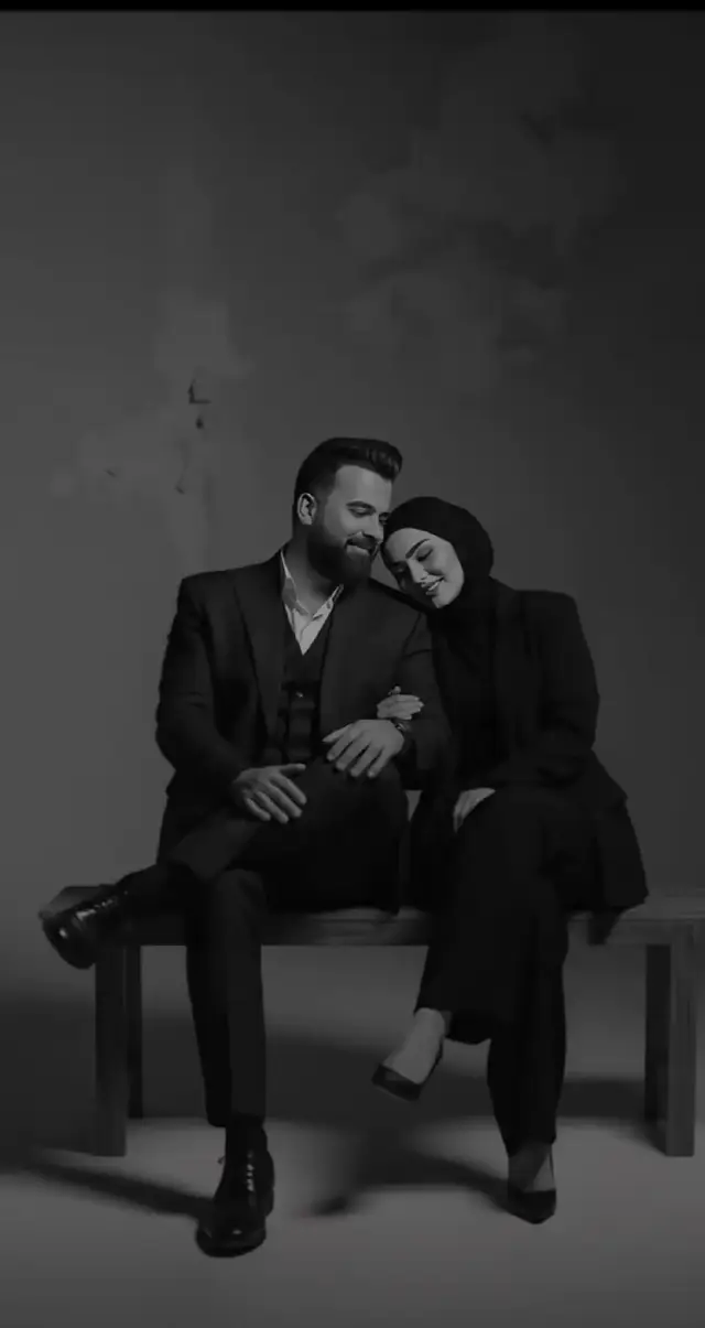 Okay, I will edit the image to a black and white studio photo showing a young woman sitting casually on a metal folding chair with her legs crossed. She is wearing a black blazer, wide black fabric trousers, and black high-heeled shoes, with a black hijab covering her head and neck completely. The lighting is soft and simple, creating a steady shadow and mood. Next to her, a man wearing a nice suit sits on a chair, looking at her. He is slightly leaning with one hand on the back of his chair, and his face is turned to the side. The background is simple, with minimalist and modern aesthetics, without altering any facial features. Here is the image