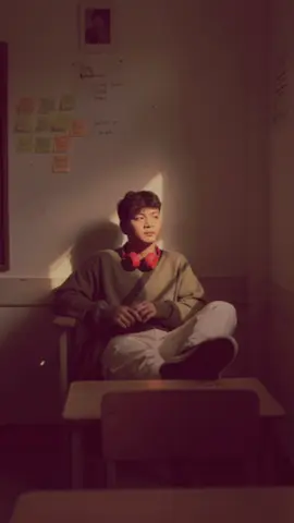 promptnya : Gemini AI prompt ➡️Original Face, cinematic portrait with a somber feel features a young Asian man in a dimly lit, vintage classroom. The camera is shot from a distance, with a small section of the chair's back visible in front, as if the photographer was taking the portrait discreetly or in a quiet moment. Soft, golden-yellow lighting reflects from a small window or gap in the right wall, forming diagonal lines of light that fall directly on the man's face, creating a dramatic and emotional contrast. The man sits alone in a chair, his feet resting on the desk, with a relaxed and cool expression, as if lost in thought. He has messy, yet natural Korean-style Comma-style hair. He wears an oversized army green Sweater, cream-colored cargo pants, Converse sneakers, and red headphones. He has a bag hanging from the chair, but it blends harmoniously with the warm light surrounding him. Behind him is an off-white wall with sticky notes labeled 
