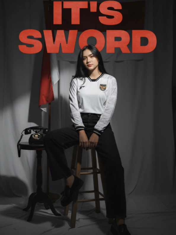 Buatkan Sebuah potret studio seorang wanita pada gambar ( pertahankan keaslian wajah ) Ia menggunakan Jersey Timnas indonesia away lengan panjang putih ( masukan gambar), dengan mengenakan celana jeans hitam panjang longgar dan sepatu Loafer docmart slop Buat dengan nuansa kasual dan retro. Pencahayaan dramatis, dengan sebagian wajah dan tubuhnya diterangi oleh cahaya berbentuk segitiga tajam dari samping, menciptakan bayangan yang kuat dan kontras yang tinggi. Latar belakangnya tirai warna putih khas studio foto dan gelap, dengan bayangan yang tidak jelas terlihat di sisi kanan. Pose sedang duduk di kursi kayu tinggi vintage dengan kaki menyilang, gaya casual dan cool seperti model internasional  Juga terdapat sebuah meja dan telepon jadul dan di belakang terdapat bendera merah mutih digantung. #promptgemini #ai  #geminiai #promptai 