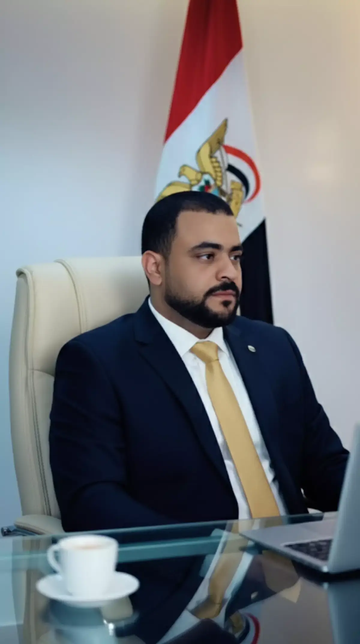 A cinematic portrait of the same إليك الوصف voung man from the providea photo, I keeping his real face and natural features unchanged. He is sitting on a modern executive chair upholstered in cream leather positioned behind a glass-top desk with chrome edges. The man wears a dark navy suit with a golden tie looking to the .side with a serious and confidentt expression flag is proudly بدون نجمة Behind him the Yemen displayed. The scene is lit with soft cinematic lighting to highlight his facial details the glossy reflections of the glasst desk, I and the textures of the suit A blurred laptopr and a coffee cup are placed in the foreground to add realism and   .#لايك__explore___ #المرتاح711 @المرتاح711 الحساب الأحتياطي @المرتاح711 denth