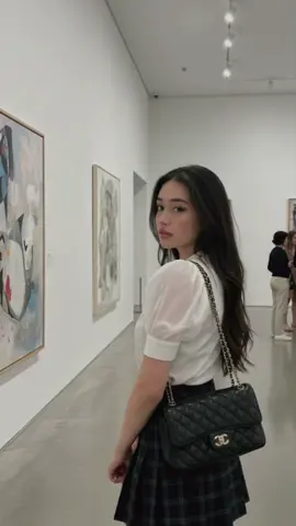 MUSEUM GIRL PROMPT: using this face, do not alter or enhance the face in any way:  Create a young girl,  taken inside a spacious and brightly lit art gallery. The central figure is a young woman with long, dark hair. She is captured from a side angle as she looks over her shoulder towards a painting on the wall to her left. Her expression is calm and contemplative. She is wearing a stylish outfit consisting of a white blouse with sheer, puffy short sleeves and a dark, pleated plaid skirt. A classic black quilted handbag with a chain strap is slung over her shoulder. The gallery setting is characterized by high, white walls, polished light-colored floors, and professional track lighting on the ceiling. Several other framed artworks are visible in the background, along with a few other museum patrons quietly observing the pieces. The overall atmosphere of the image is quiet, sophisticated, and serene. #gemini #ai #prompt 