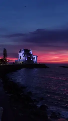 How early is too early? In Constanța, winter answers with a hush, an indigo vastness where sea and sky are indistinguishable conspirators, and a filament of pink-orange trembles along the horizon like a pulse under porcelain skin. The Black Sea does not blaze at first light, it smolders. The Casino, that Art Nouveau reverie inaugurated in 1910, stands on its promontory like a dowager queen contemplating eternity, its silhouette cut from shadow, its windows still holding the last secrets of the night. This shore was once Tomis, a Greek colony where traders counted coins and exiles counted regrets, most famously the poet Ovid, banished here by Augustus to the edge of the known world. Imagine him watching a dawn like this, the Pontus Euxinus stretching iron-blue before him, winter winds sharpening his verses. The sea itself is a peculiar alchemy, less saline than the Mediterranean, layered and stratified, its deeper waters almost devoid of oxygen, preserving wrecks and relics in a kind of submerged time capsule. And above it all, the Casino rose in the early twentieth century as a declaration that Romania belonged to the grand conversation of Europe, a flourish of curves and ornament facing a horizon that has swallowed empires without ceremony. Winter strips the coast of chatter and leaves only essence. The sea becomes philosophy made liquid, austere and exacting, a cold mirror that refuses vanity. That narrow band of fire at the horizon feels less like sunrise and more like revelation, a reminder that beauty does not clamor, it endures. Standing there, wrapped in salt air and silence, you sense how small you are and how magnificently alive. Do you seek the sea when it is gentle, or when it is severe?  Video by @vvh_photography [ Constanta Casino, Black Sea Winter, Art Nouveau Romania, Tomis Ancient City, Ovid Exile, Romanian Seaside, Dobrogea History, Black Sea Coast, Winter Sunrise, Eastern Europe Travel, Historic Constanta, Pontus Euxinus, Sunrise Over Sea, Coastal Architecture, Romanian Landmarks ] #romania #travel #constanta #blacksea