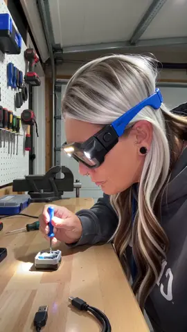 @🛒✨DailyFindsWithManda💜🛍️  Create an ultra-realistic 20-second TikTok product demonstration video in vertical 9:16 format using the uploaded JOYTUTUS Rechargeable LED Headlamp Glasses product image as the exact reference. A friendly, confident American woman is wearing the EXACT glasses from the reference image. The glasses must have black rectangular frames, bright blue arms, a long horizontal LED light across the upper front frame, and two round LED spotlights positioned on the outer sides. ACTION: The woman is standing in a garage at a workbench, wearing the glasses while working on a small household project. She looks directly at the camera and talks naturally while demonstrating the product. She switches on the lights, illuminating her work area, then waves her hand near the sensor to demonstrate the hands-free light control. She continues working with both hands while wearing the glasses, showing how practical they are. Keep the camera moving smoothly in ONE continuous shot. No scene changes or cuts. SPOKEN SCRIPT — Natural American Female Voice: 